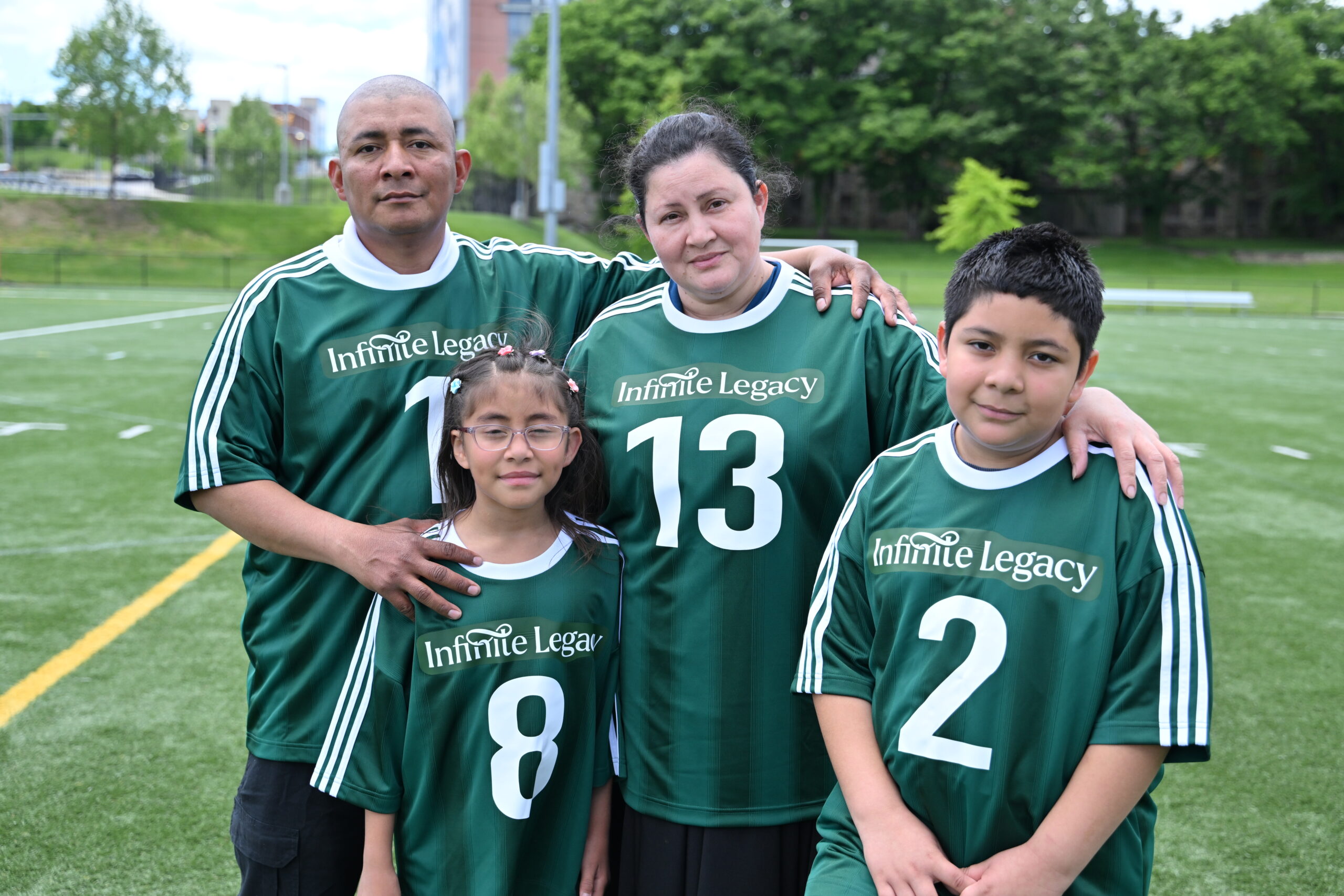 Emily Bardales, receptora de un trasplante de corazón, junto a su familia, que lleva camisetas de fútbol verdes, posan juntos en un campo de césped artificial, abrazados, para una foto de grupo.