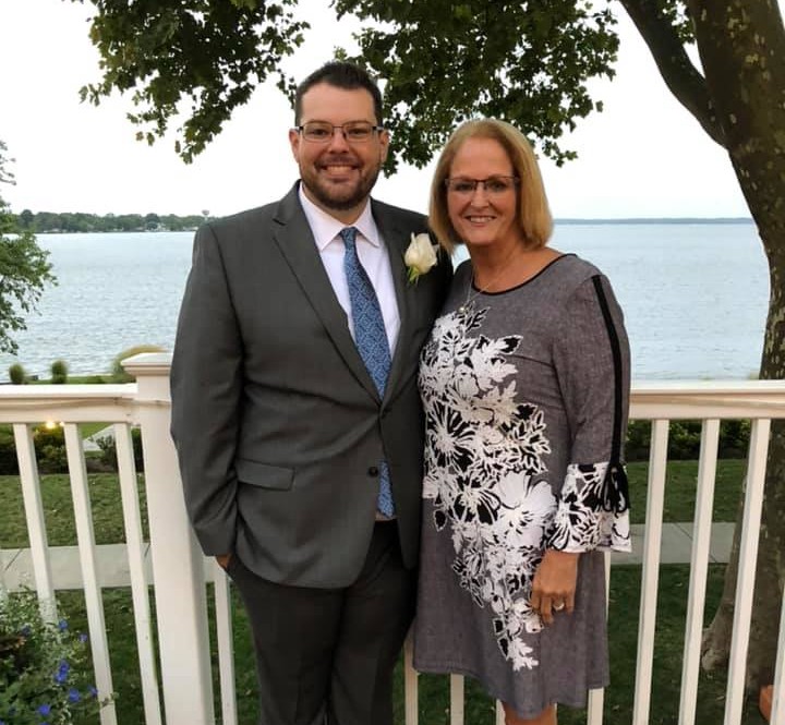 A couple posing outdoors by a lake; man in a gray suit with a boutonniere and a woman in a gray floral dress smile at the camera on a white railing backdrop.