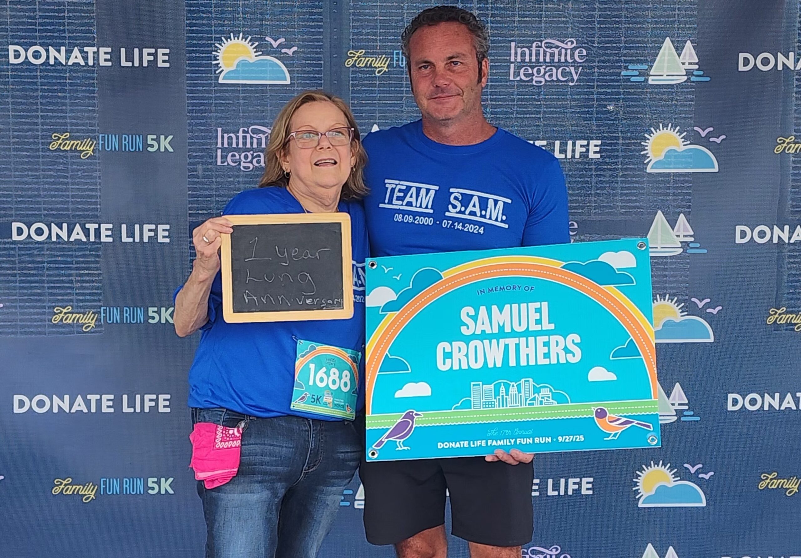Two people in blue shirts posing at a Donate Life event backdrop, holding a small chalkboard and a bright commemorative sign for Samuel Crowthers.