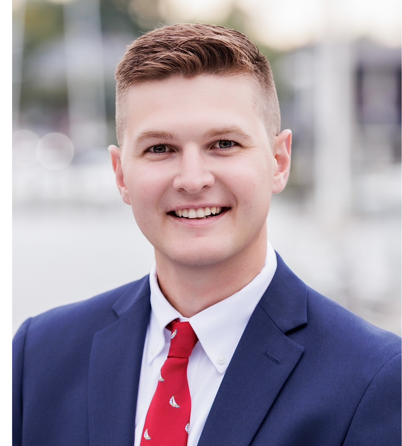 Smiling young man in a navy suit and red tie, outdoor professional headshot.