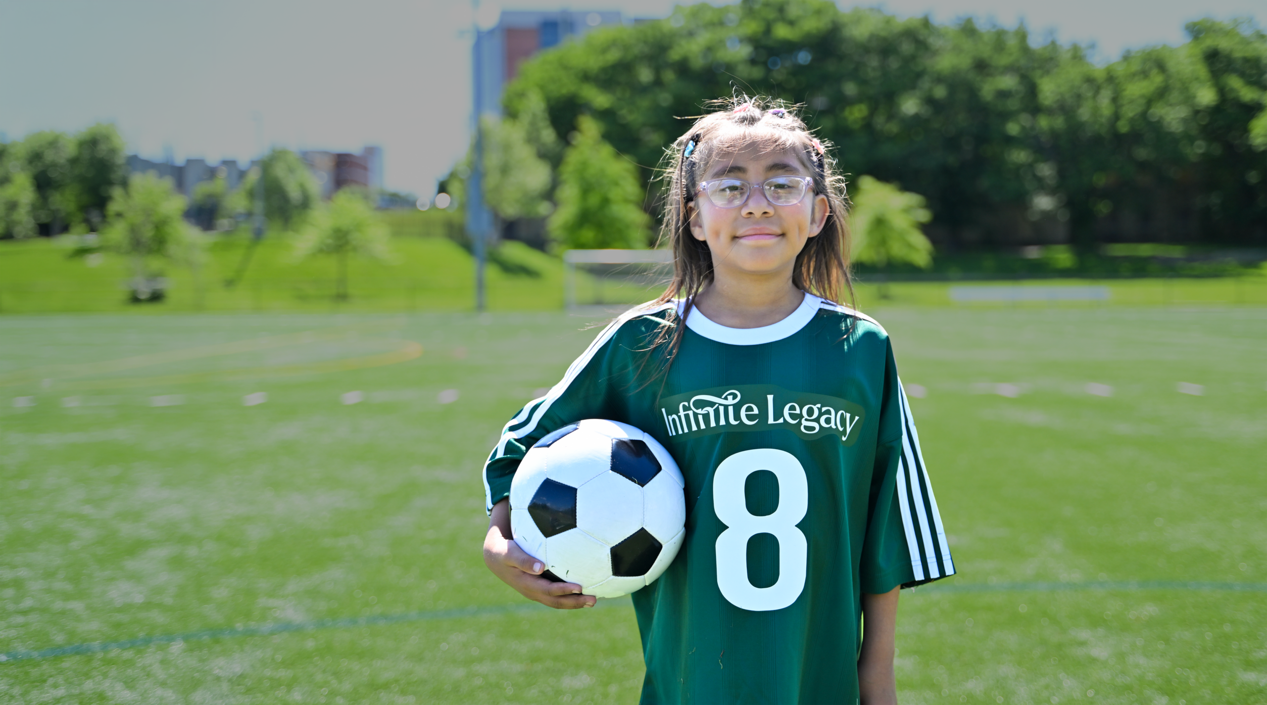 Emily Bardales, a pediatric heart recipient wears a green Infinite Legacy soccer jersey and holds a soccer ball on a sunny outdoor field.