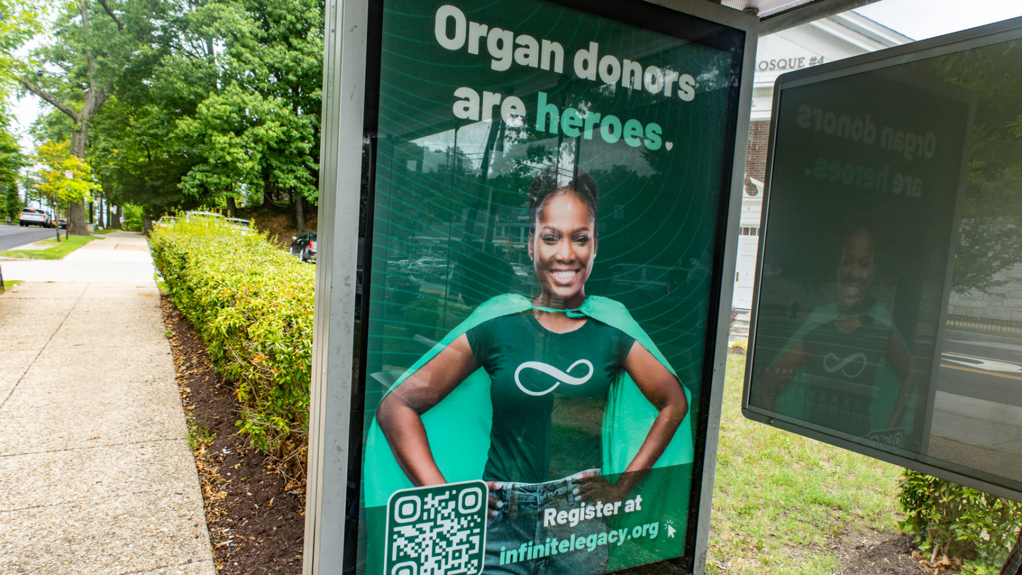 Bus-stop poster featuring a smiling woman in a green cape; slogan 'Organ donors are heroes' with a QR code visible.