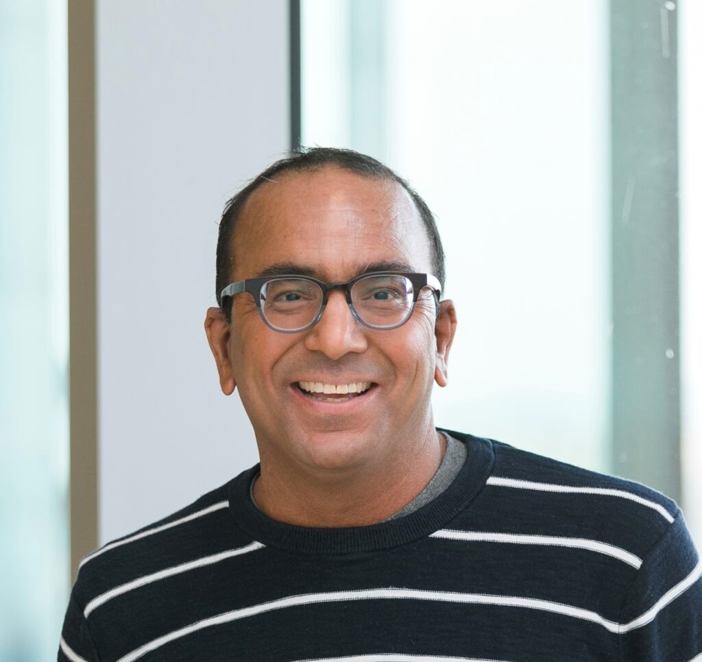 Hombre sonriente con gafas y una camiseta a rayas blancas y negras en un interior, con el encuadre a la altura del pecho.