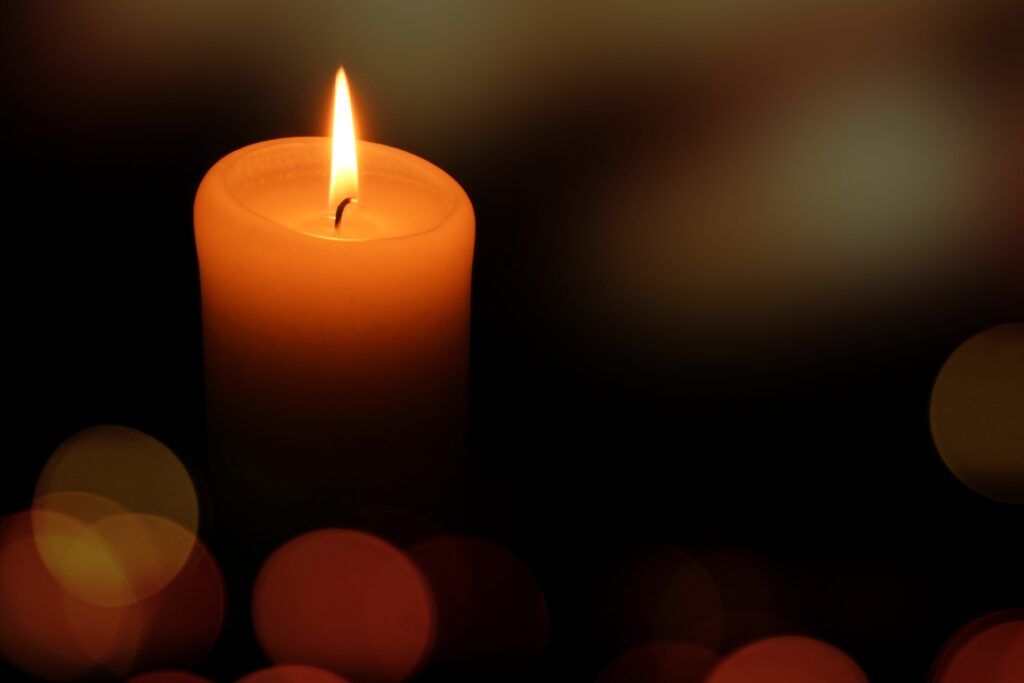 Una vela cilíndrica naranja encendida con una llama alta en una habitación a oscuras, con luces cálidas y difuminadas al fondo.