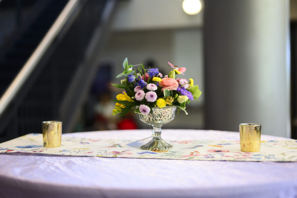 Ramo de flores de colores en un jarrón de cristal con pedestal, sobre un mantel estampado con un fondo difuminado.